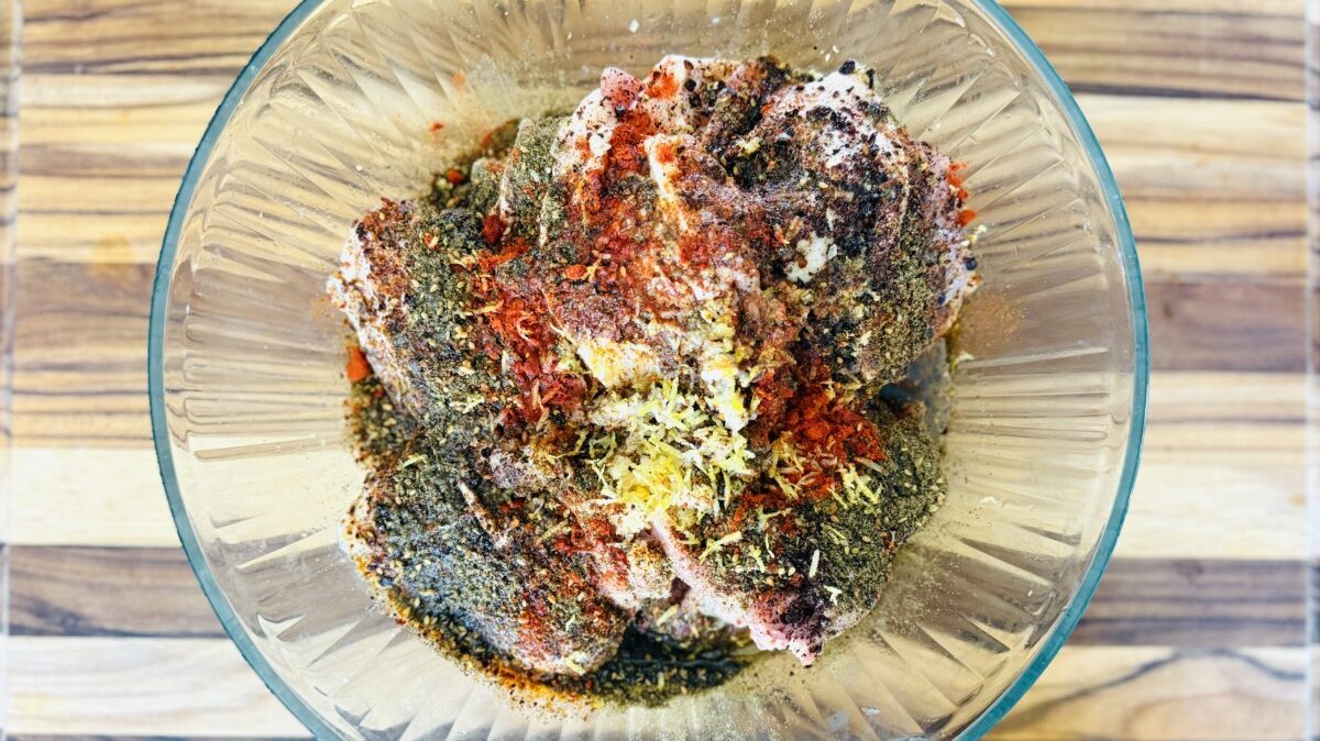 Raw chicken thighs in a mixing bowl topped with za’atar, sumac, garlic, lemon, olive oil, and spices, ready to be mixed.