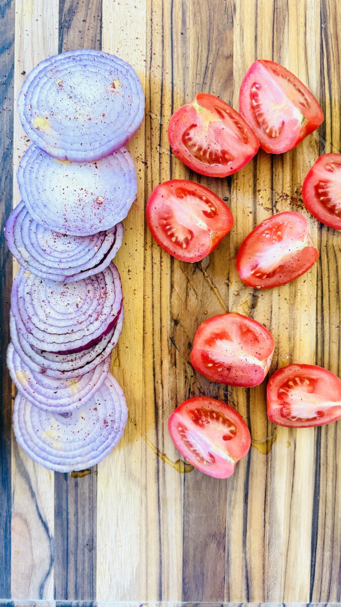 Halved ripe tomatoes and thick-cut red onion wedges arranged on a cutting board, ready for grilling.