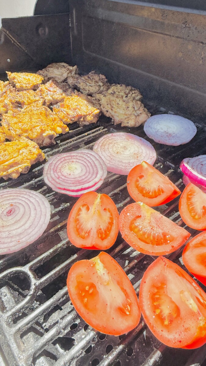 Halved tomatoes and onion wedges placed on the grill next to chicken thighs during final stage of cooking