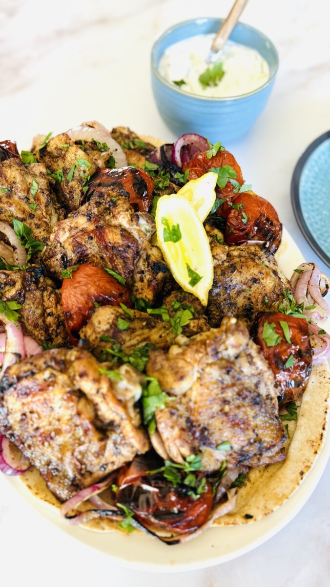 Large platter of grilled za’atar chicken thighs with roasted tomatoes and onions, served with a bowl of yogurt sauce in the background.