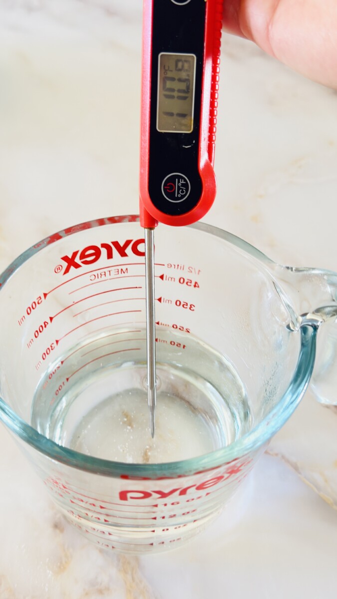 Using a thermometer to test warm water between 100–110°F for pita bread dough.