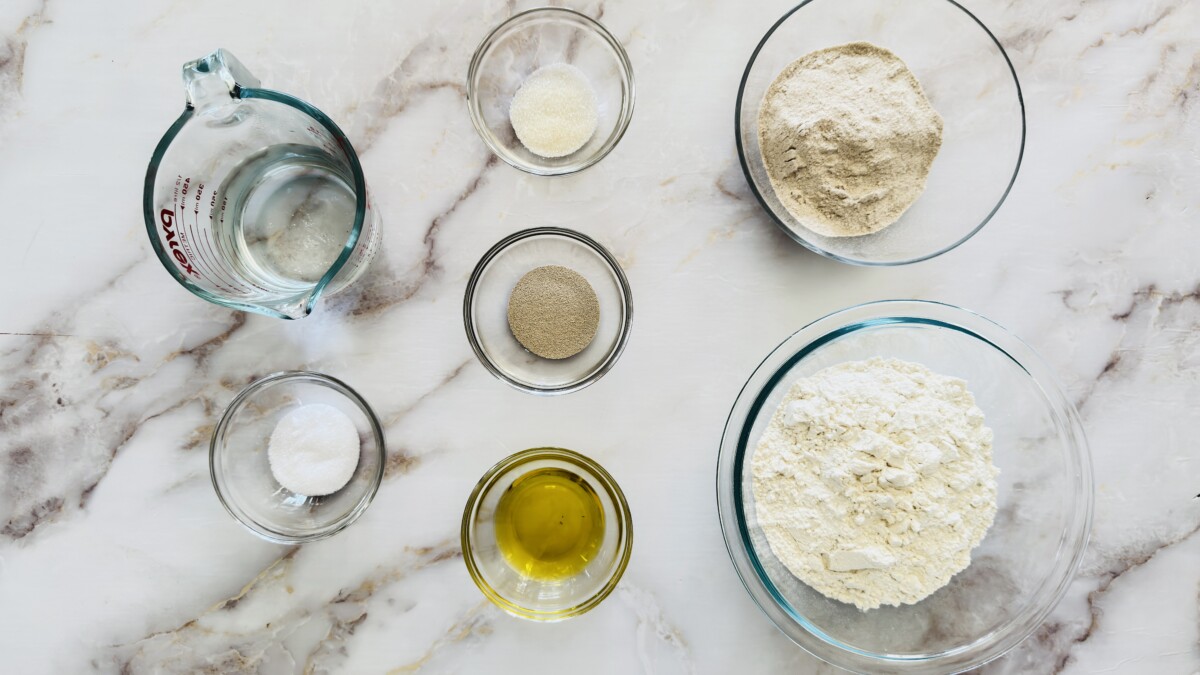 alt="Top-down view of pita bread ingredients on a wooden surface, including flour, olive oil, yeast, salt, and water in small bowls" 