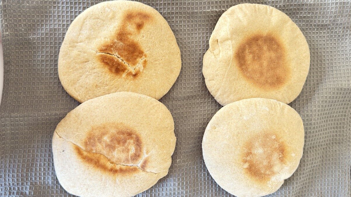 alt="Golden puffed pita bread just out of the oven, resting on a striped kitchen towel to cool