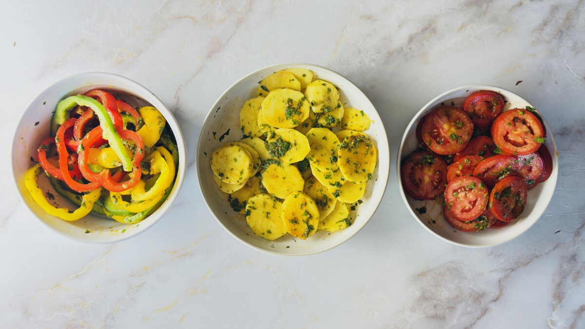 Bowls of sliced potatoes, tomatoes, and colorful bell peppers coated with Moroccan chermoula marinade — vegetables ready to layer in the fish tagine.