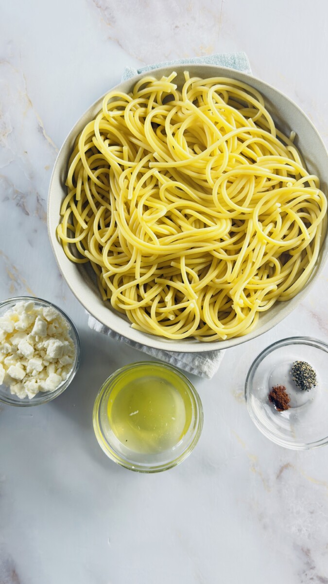 Cooked pasta in a bowl with feta, egg whites, and spices on the side — ready to be mixed for the Greek Pastitsio pasta layer.