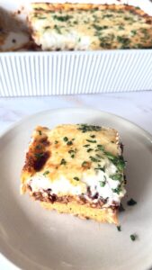 A slice of homemade Greek Pastitsio topped with golden béchamel and fresh parsley, served on a plate with the remaining casserole in the background.