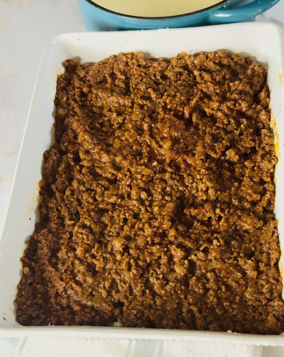 Spreading rich meat sauce evenly over the pasta layer to build the Greek Pastitsio before adding béchamel.