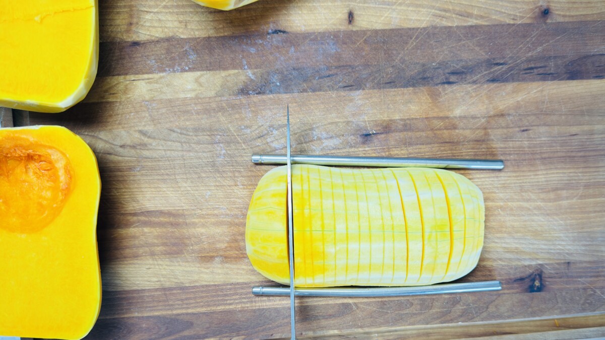Butternut squash half between two chopsticks being sliced hasselback-style with a knife