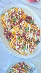 Hasselback butternut squash with pomegranate seeds, pine nuts, crispy pancetta, and tahini drizzle on serving platter with side plate of sliced portions
