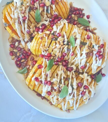 Hasselback butternut squash with pomegranate seeds, pine nuts, crispy pancetta, and tahini drizzle on serving platter with side plate of sliced portions