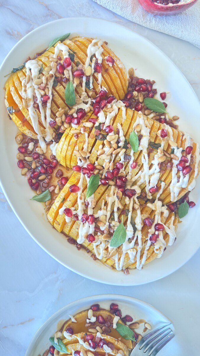 Hasselback butternut squash with pomegranate seeds, pine nuts, crispy pancetta, and tahini drizzle on serving platter with side plate of sliced portions