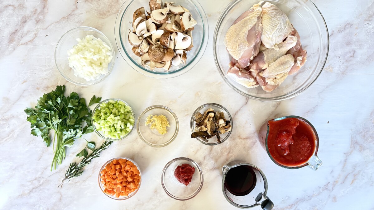 Ingredients for chicken cacciatore including whole chicken pieces, dried porcini mushrooms, cremini mushrooms, red wine, crushed tomatoes, carrots, celery, onions, and fresh herbs on a white surface