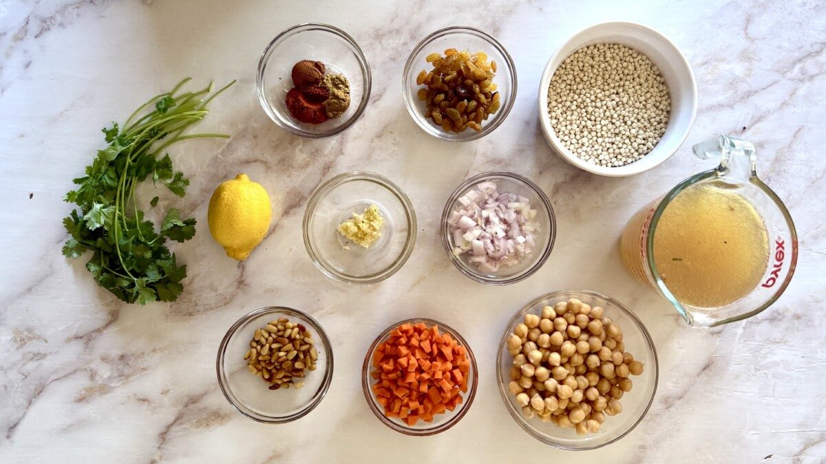 Ingredients for pearl couscous recipe including chickpeas, carrots, golden raisins, warm spices, and fresh herbs on a white surface