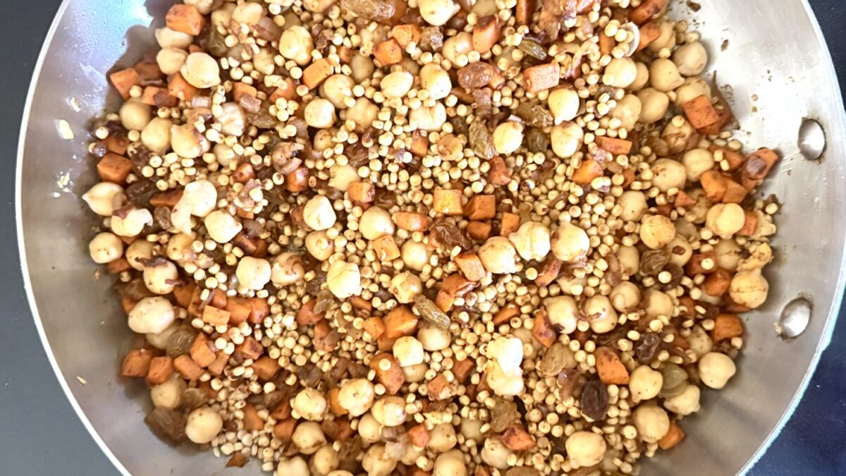 Pearl couscous being added to pan with chickpeas, vegetables, and warm spices
