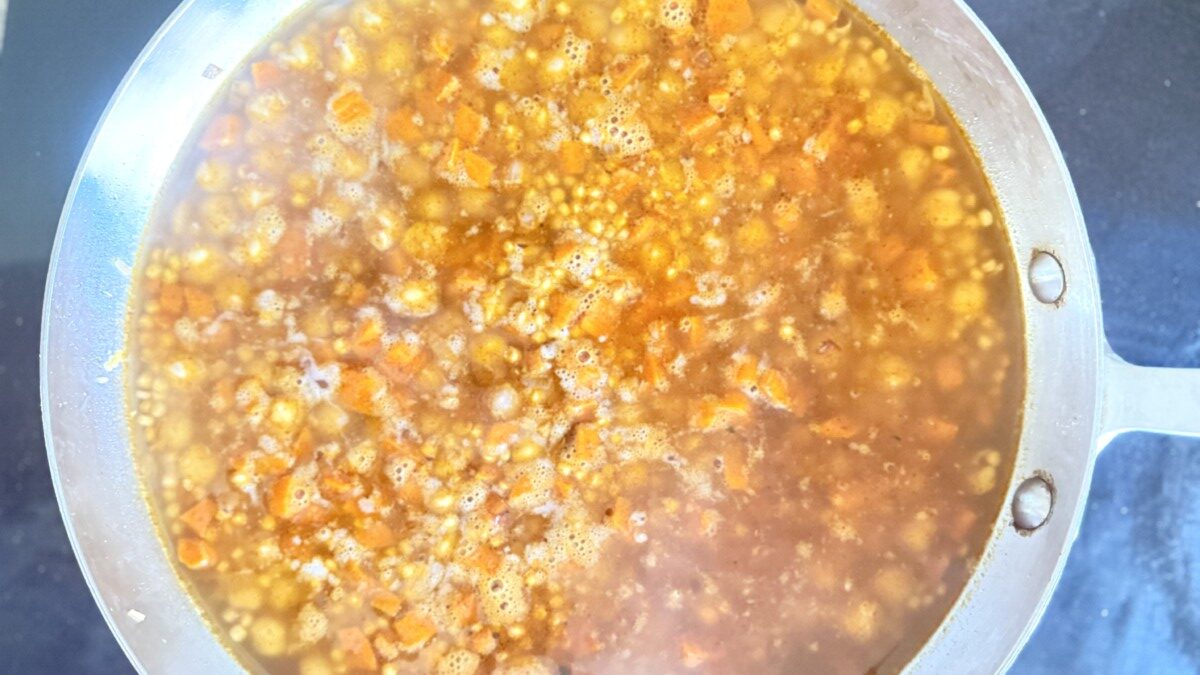 PHOTO 7: ADDING BROTH Alt Text: Vegetable broth being poured into pan with pearl couscous, chickpeas, and vegetables ready to simmer