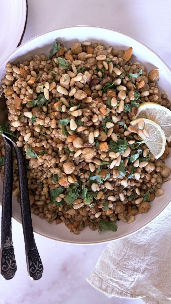 Pearl couscous with chickpeas, carrots, and golden raisins in a serving dish with two spoons and thinly sliced lemon