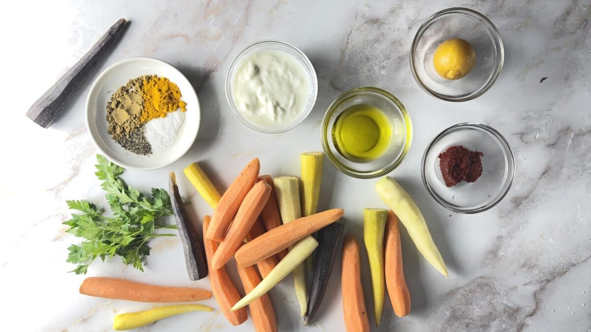 Ingredients for harissa roasted carrots including rainbow carrots, harissa paste, warm spices, preserved lemon, and Greek yogurt on a white surface