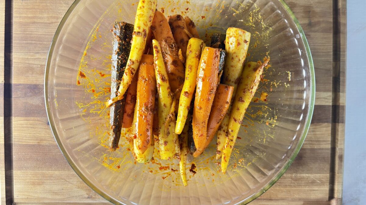 Rainbow carrots being tossed in harissa spice mixture in a bowl for roasted carrots recipe