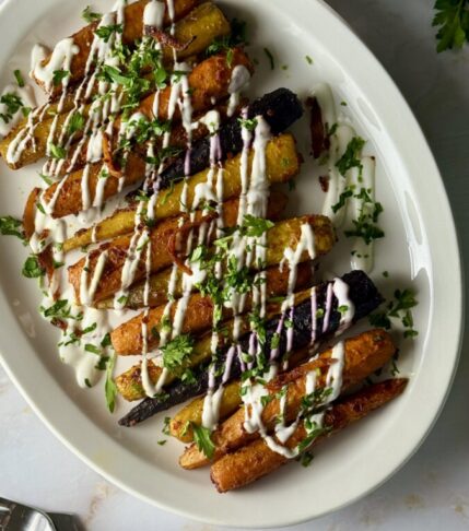 Harissa roasted carrots on a white platter drizzled with yogurt sauce and garnished with preserved lemon ribbons and fresh parsley