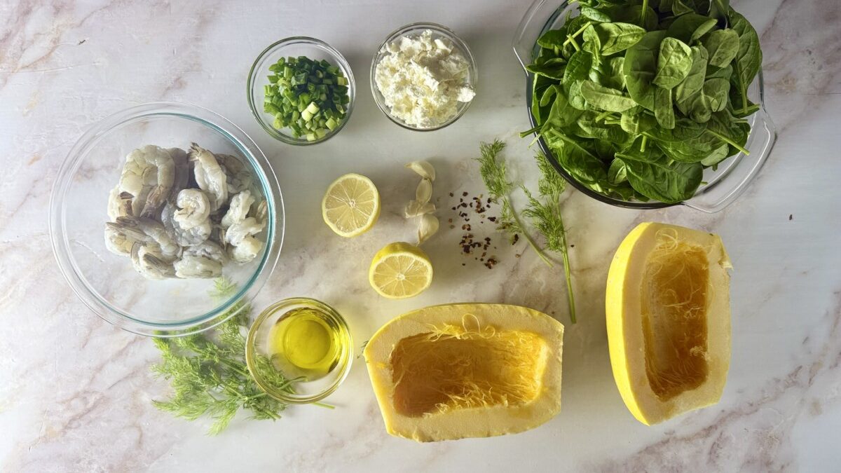 Ingredients for Mediterranean spaghetti squash recipe including halved squash, shrimp, spinach, feta cheese, lemon, dill, and garlic arranged on a white surface